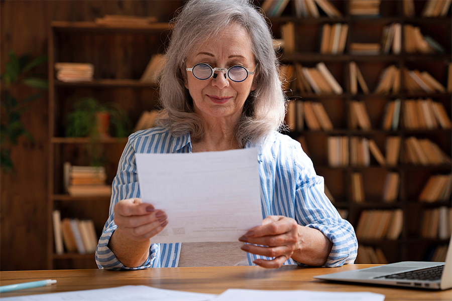 asb_lv_testamentspende_2025_freepik.jpg Ältere Frau mit grauen Haaren schaut auf ihr Testament in einem Raum mit Bücherregalen im Hintergrund.